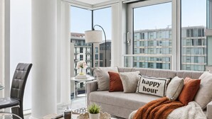 A modern living room with a sofa, a coffee table, and a chair. There is a view of buildings through large windows.