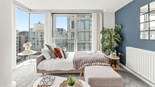 A modern bedroom with a large window, a bed with multiple pillows, a bedside table with a lamp, and a potted plant.