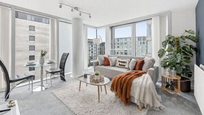 A modern living room with a glass dining table, a sofa with decorative pillows, a large plant, and a view of the cityscape through large windows.