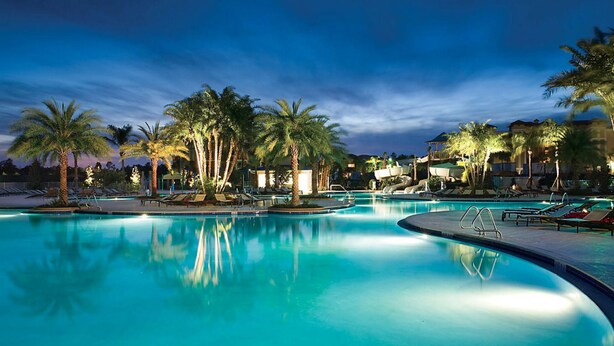A large outdoor swimming pool surrounded by palm trees and lounge chairs, with a building in the background. - Adventure Park Vacation 1 BR Condo with Tropical Pool (Orlando)