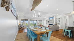 A spacious dining area with a wooden table and blue chairs, a kitchen with white cabinets, and a living area with a gray sofa. - Bonny Blue:  Gorgeous SGI Home, Pet Friendly, Private Pool,  Sleeps 14 (St. George Island)