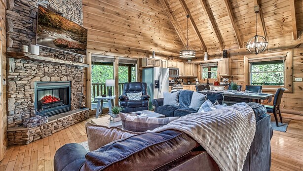 A cozy cabin interior with a stone fireplace, wooden ceiling, and a dining area.