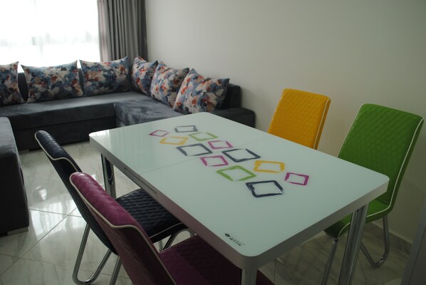 Un salon moderne avec une table blanche, des chaises colorées, un canapé gris agrémenté de coussins à motifs floraux et une grande fenêtre avec des rideaux.