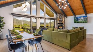 A spacious living area with a wooden ceiling, large windows offering a view of mountains, a green sofa, and a dining table set for four. - 🌙 Crescent Chalet - Hot Tub, Views, Sauna! (Oakhurst)