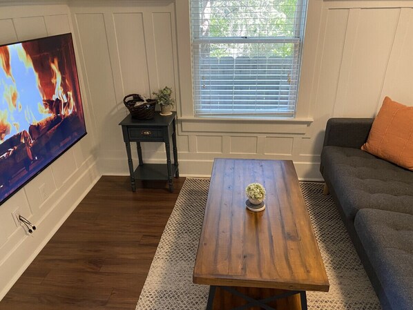 A living room with a wooden coffee table, a TV mounted on the wall, a window with blinds, a dark-colored sofa, and a small side table with a plant.