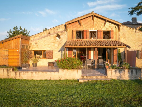 Une maison en pierre avec un toit en tuiles, des volets en bois et un patio couvert doté d’un espace salon extérieur.