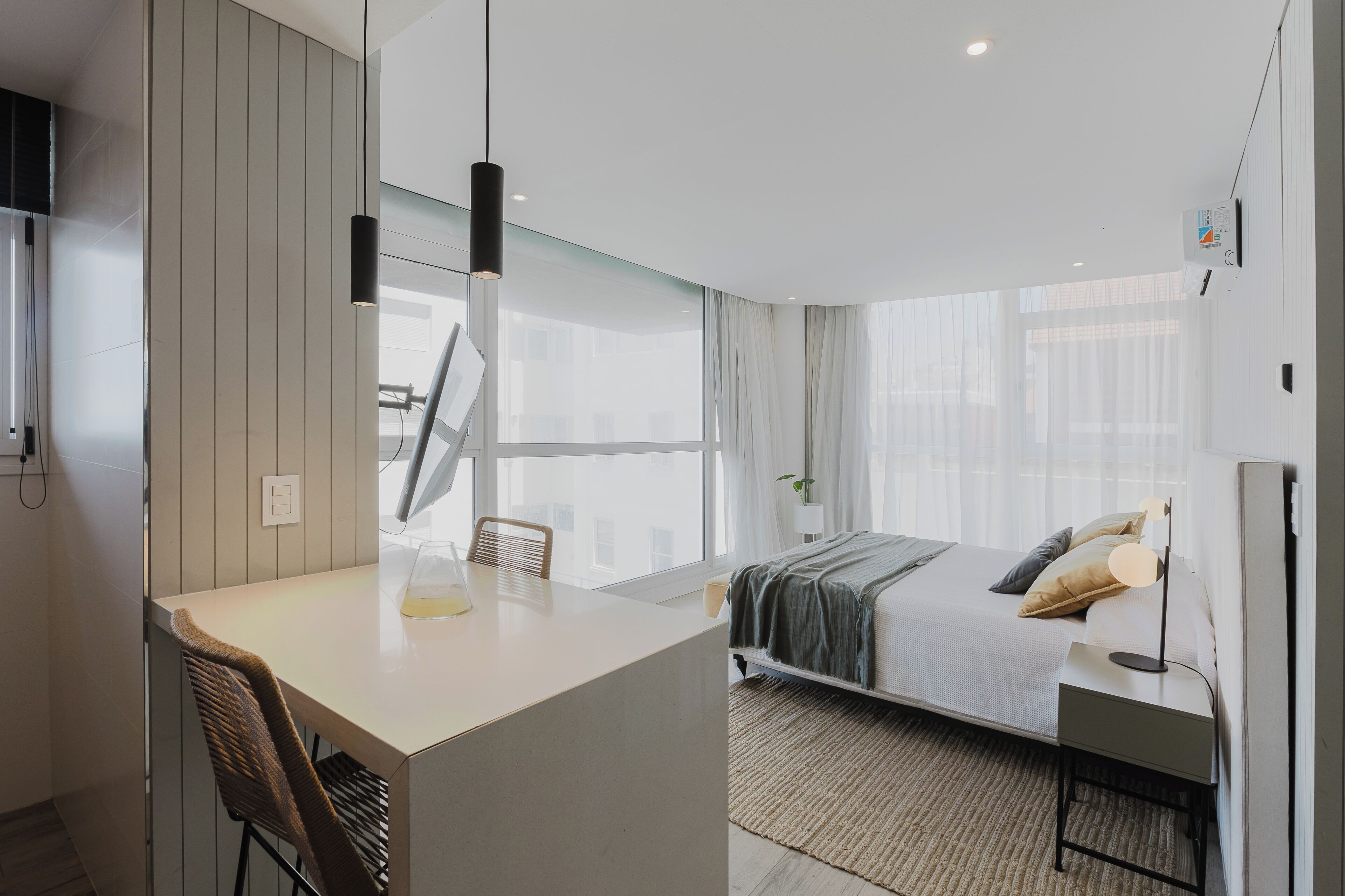 A modern hotel room with a large bed, a desk, and a chair. There is a window with curtains, a bedside table with a lamp, and a wall-mounted TV.