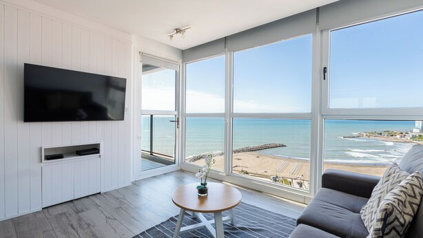 A modern living room with a large TV, a round table with a plant, and a sofa with a patterned pillow. The room has large windows offering a view of the beach and ocean. - Residencias Roque Suarez (Mar del Plata)