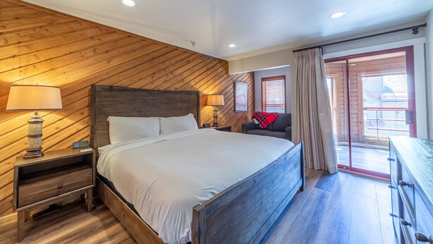 A bedroom with a wooden ceiling, a large bed, bedside tables with lamps, a dresser, and a view of the outside through a sliding glass door.