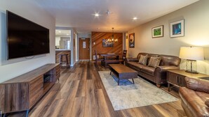 A modern living room with a large flat-screen TV, wooden flooring, a leather sofa, a coffee table, and a dining area with a wooden table and chairs.
