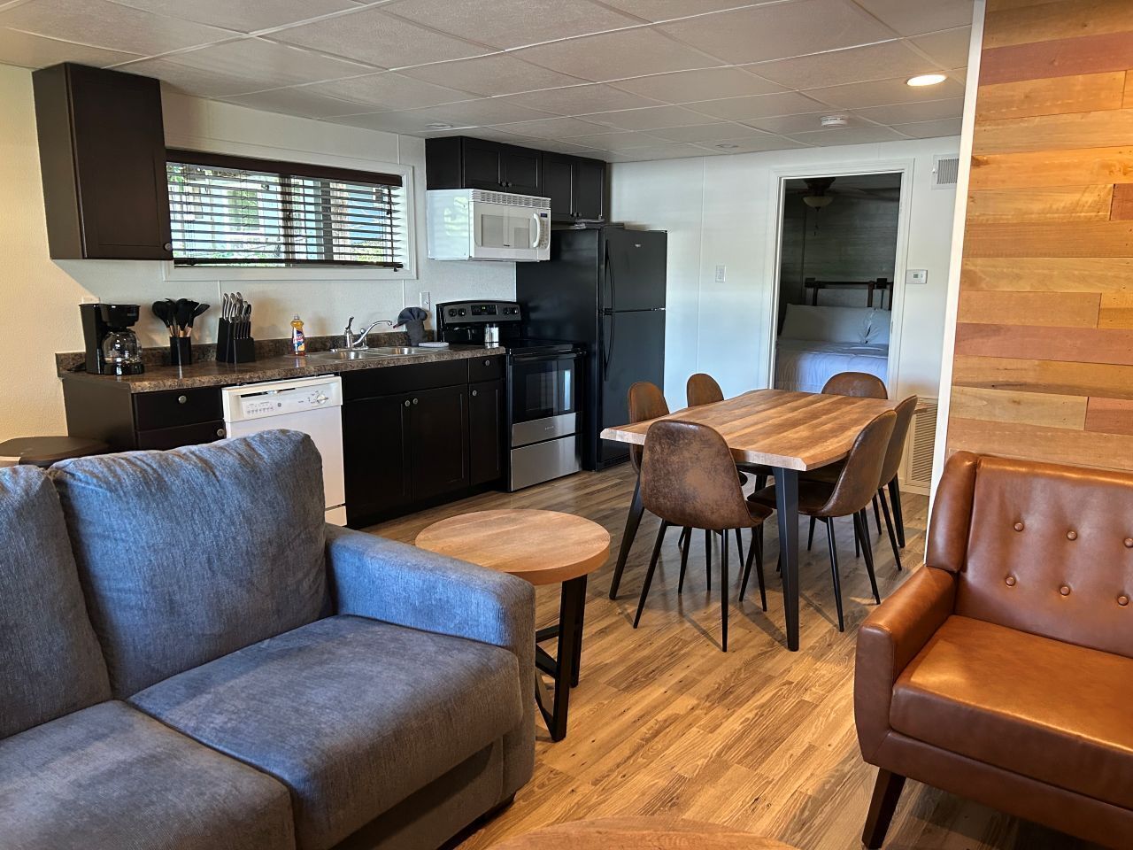 A modern kitchen with dark cabinets, a microwave, and a refrigerator. There is a dining area with a wooden table and chairs, and a gray sofa.