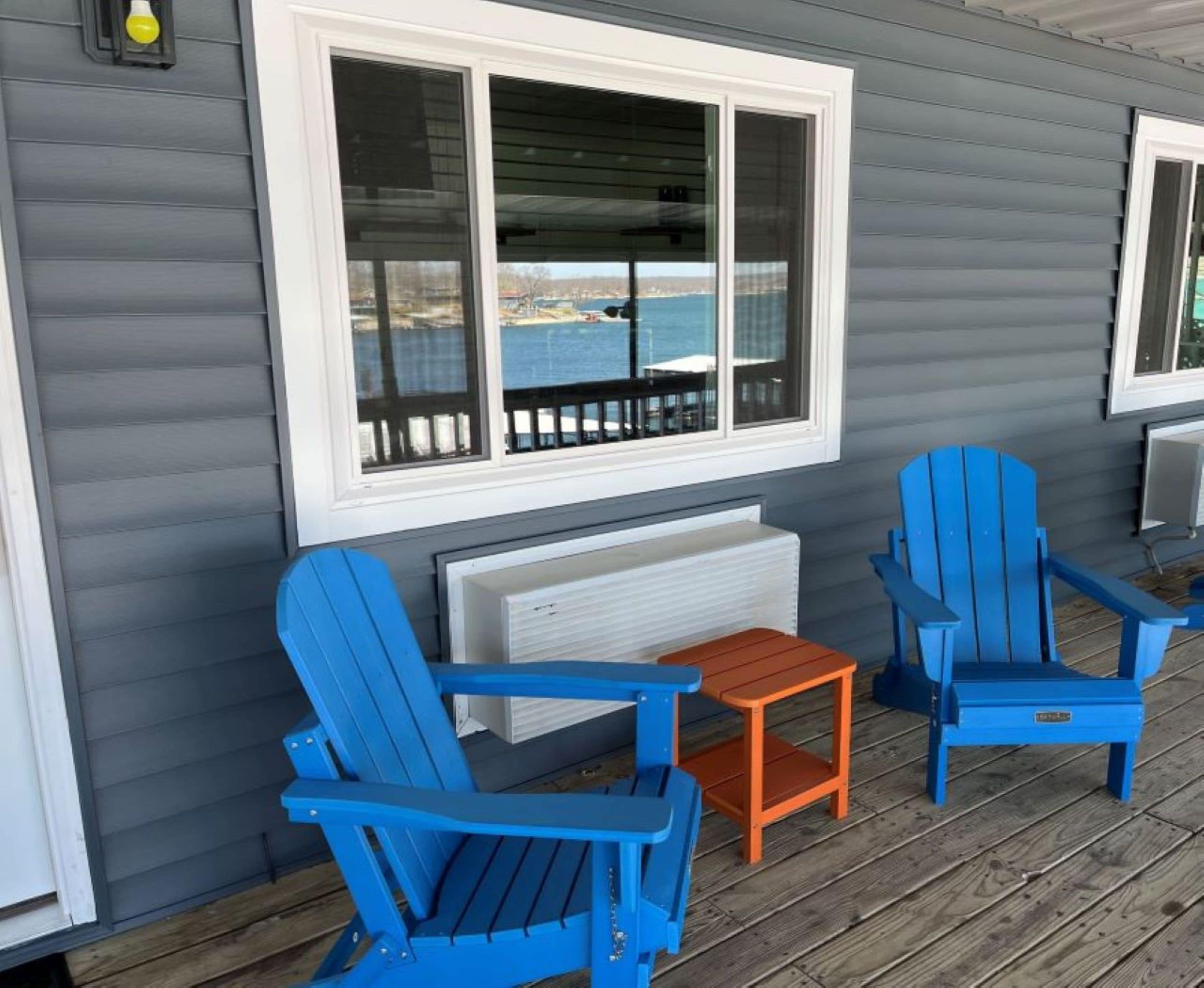 A covered porch with two blue Adirondack chairs, a small orange table, and a white radiator below a large window overlooking a body of water.