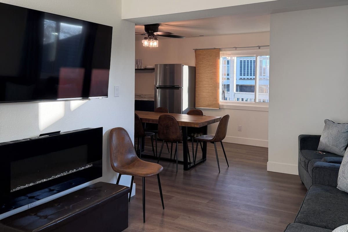 A modern living room with a flat-screen TV, a dark entertainment unit, a grey sofa, and a dining area with a wooden table and chairs.