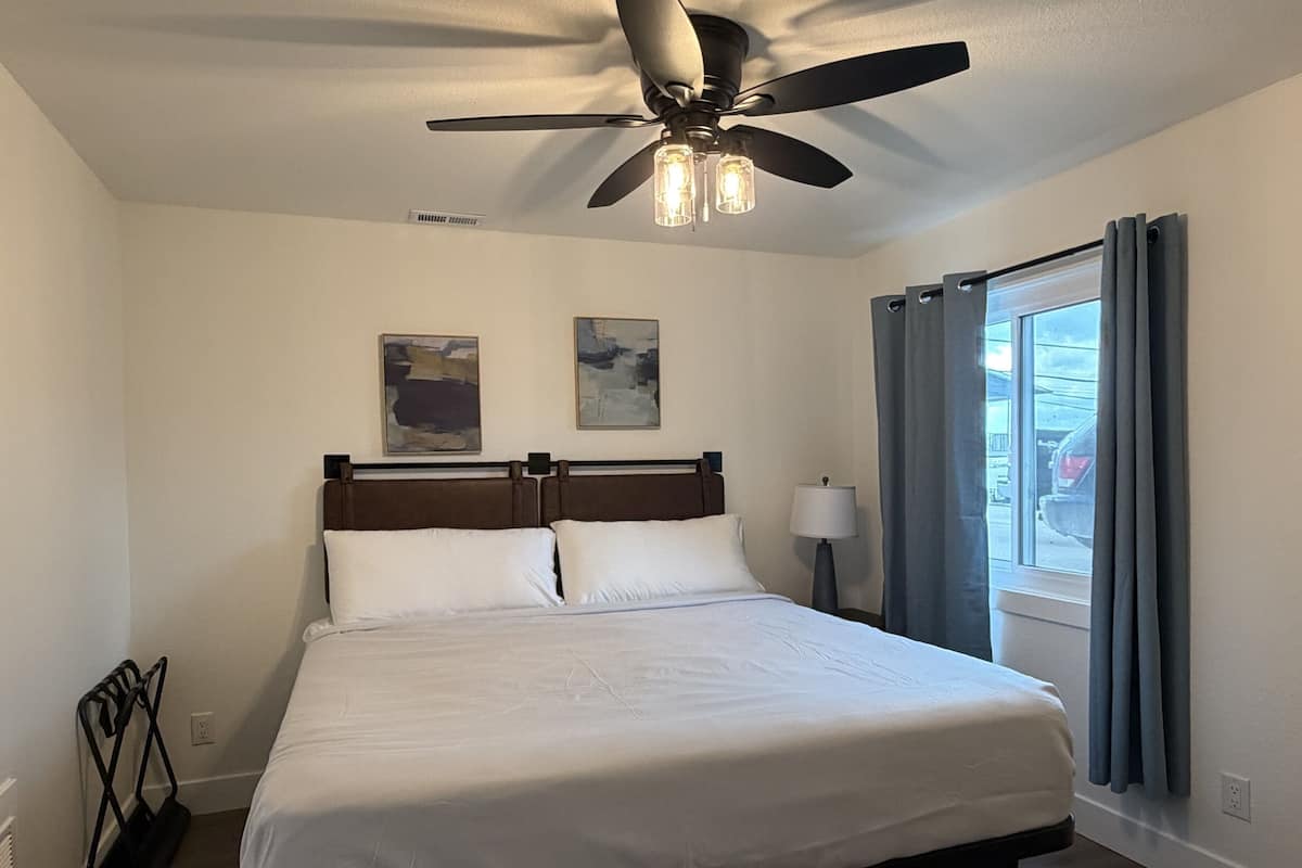 A hotel room with a bed, ceiling fan, artwork, window with curtains, and a lamp.