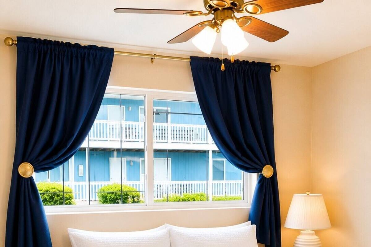 A hotel room with a bed, curtains, a ceiling fan, a nightstand with a lamp, and a view of a building outside.