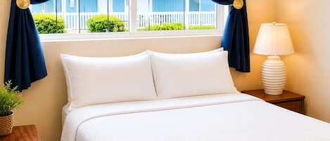 A hotel room with a bed, curtains, a ceiling fan, a nightstand with a lamp, and a view of a building outside.