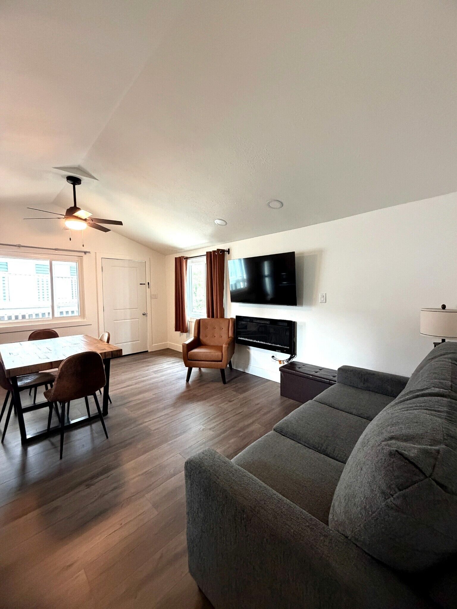 Un salon moderne avec un canapé gris, une table à manger et des chaises, un téléviseur fixé au mur et un ventilateur de plafond.