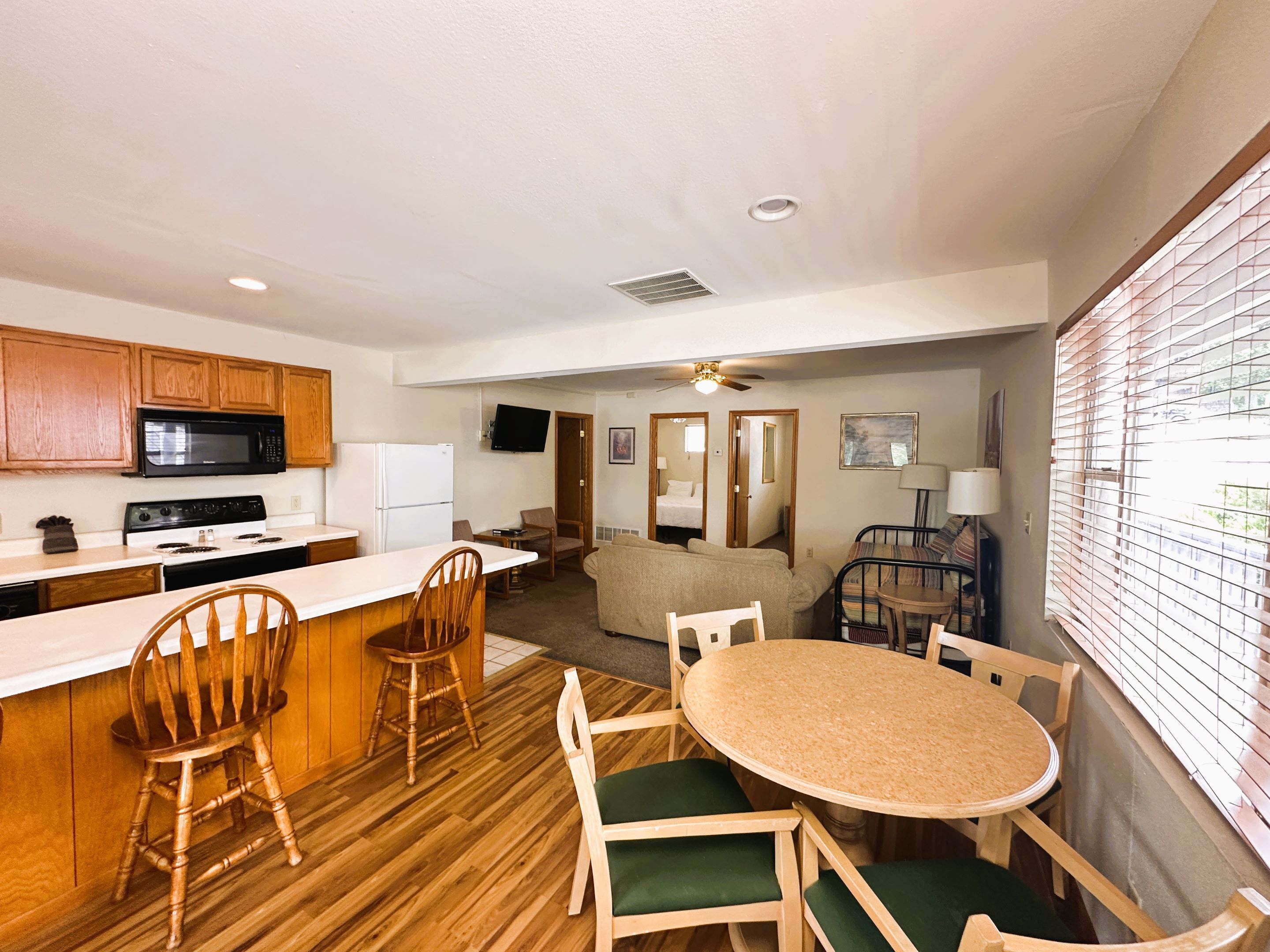 A compact kitchen with wooden cabinets, a microwave, and a refrigerator. There is a dining area with a round table and chairs, and a living area with a sofa and a TV.