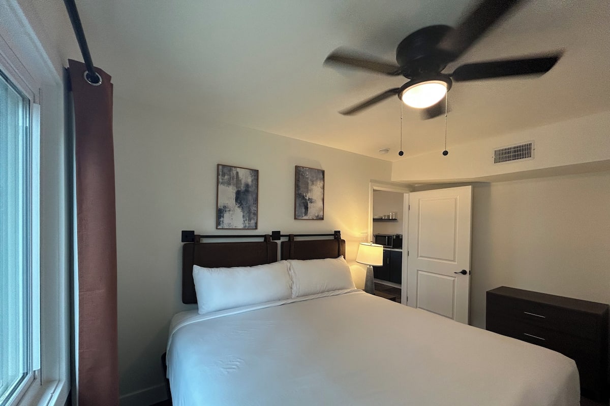 A hotel room with a bed, ceiling fan, artwork on the wall, and a door leading to a kitchenette.