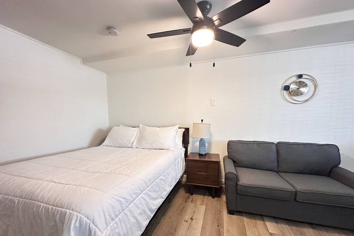 A bedroom with a bed, a gray sofa, a ceiling fan, a wall clock, and a nightstand with a lamp.