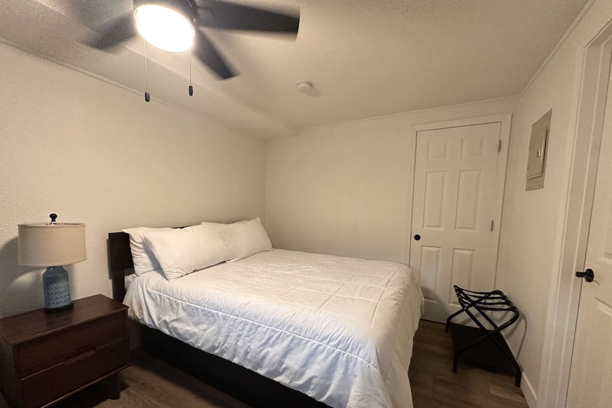 A bedroom with a bed, a nightstand, a lamp, a ceiling fan, and a door.