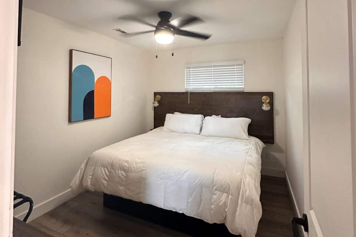 A bedroom with a bed, a ceiling fan, a wall-mounted light, and a window with blinds.