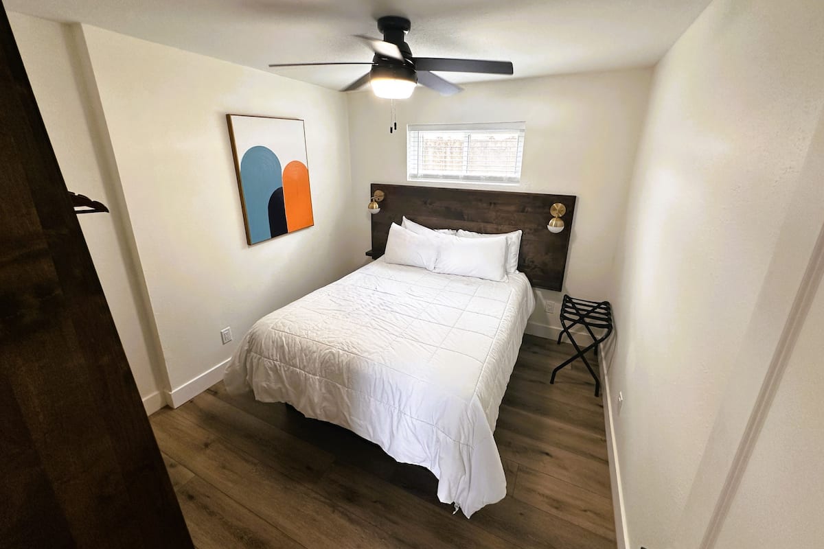 A bedroom with a bed, a ceiling fan, a window, and a wall-mounted artwork.