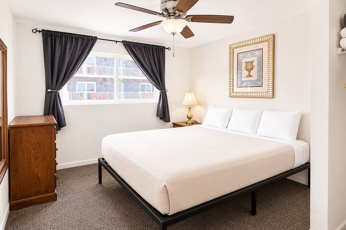 A bedroom with a bed, a nightstand, a lamp, a ceiling fan, a window with curtains, a framed picture, and a wardrobe.