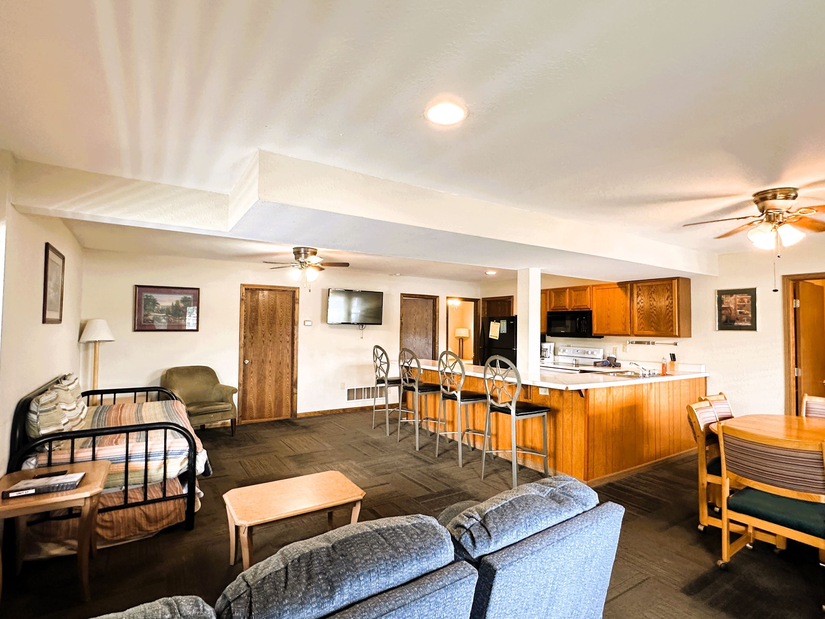 A spacious living room with a sofa, a small table, and a TV mounted on the wall. There is a kitchen area with wooden cabinets and a bar counter.