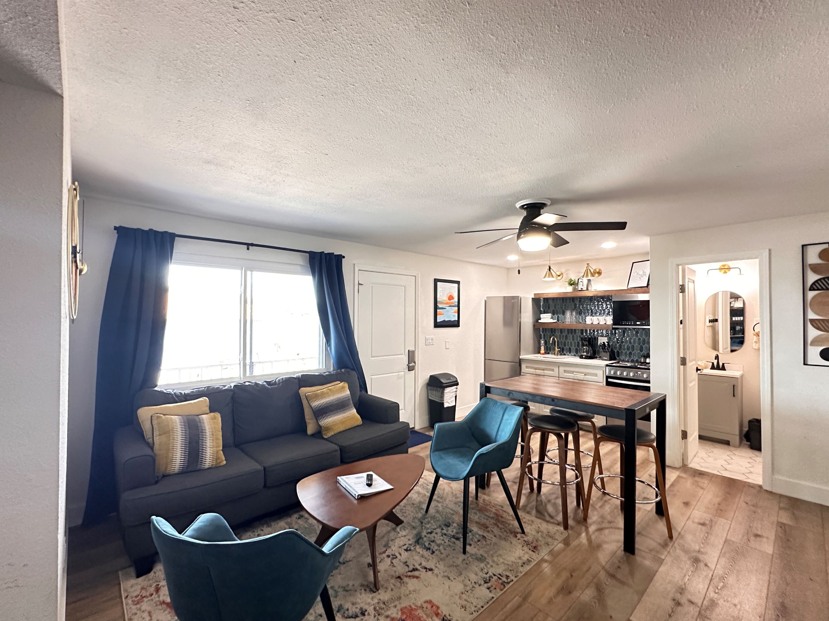 Un espace de vie compact comprenant un canapé, une table basse et un coin repas avec des chaises et une table. On peut également voir un ventilateur de plafond et une cuisine équipée d’un réfrigérateur et d’un évier.
