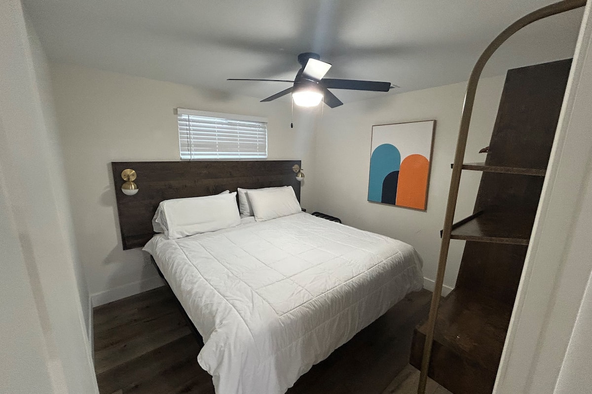 A bedroom with a bed, a ceiling fan, a window with blinds, and a wall-mounted shelf.