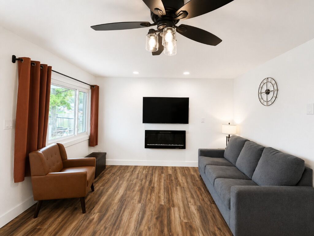 Una sala de estar moderna con un ventilador de techo, un televisor de pantalla plana montado en la pared, un sofá gris, un sillón de cuero marrón y una ventana con cortinas.