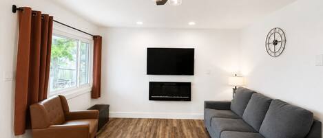 A modern living room with a ceiling fan, a flat-screen TV mounted on the wall, a gray sofa, a brown leather chair, and a window with curtains.