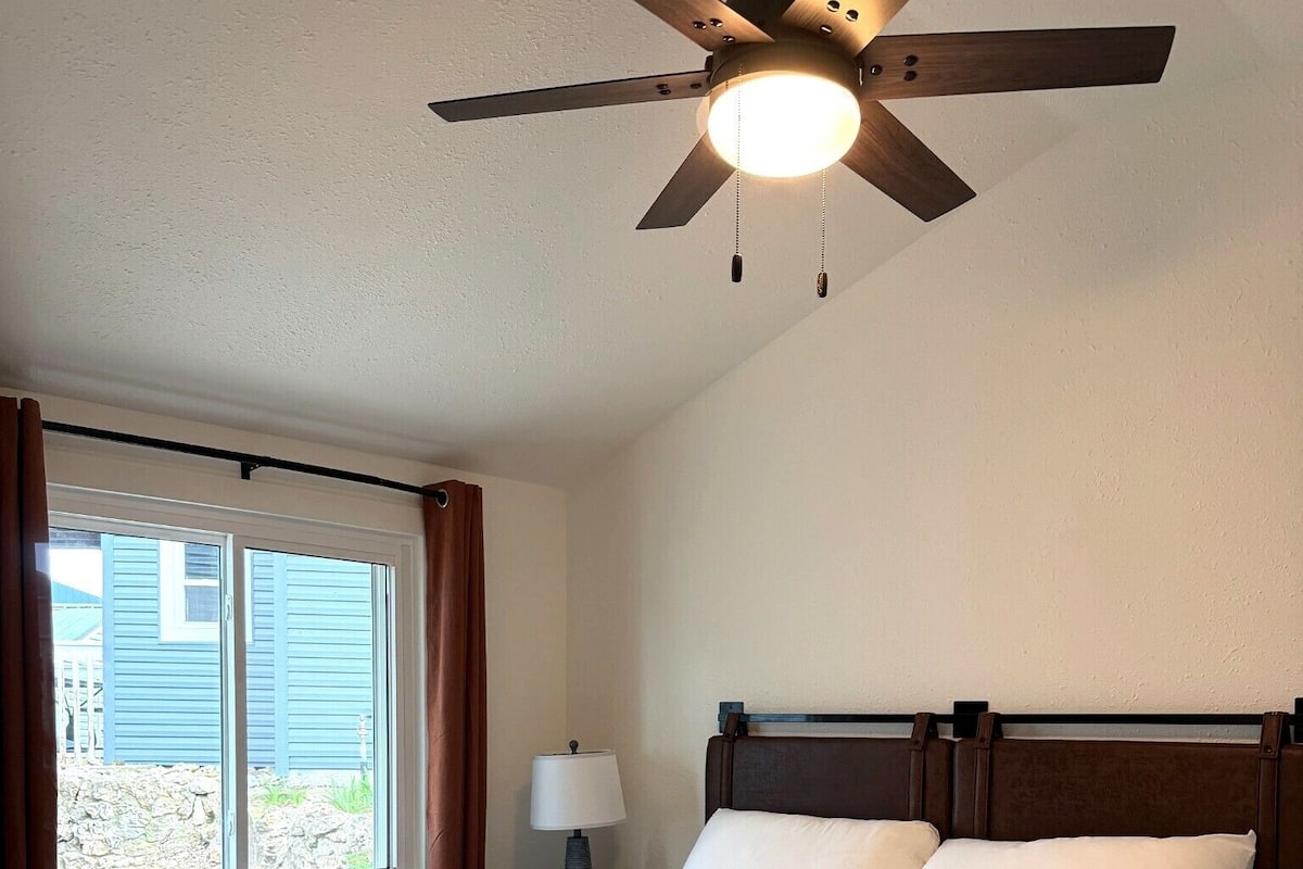 A bedroom with a bed, a ceiling fan, a window with curtains, and a lamp.