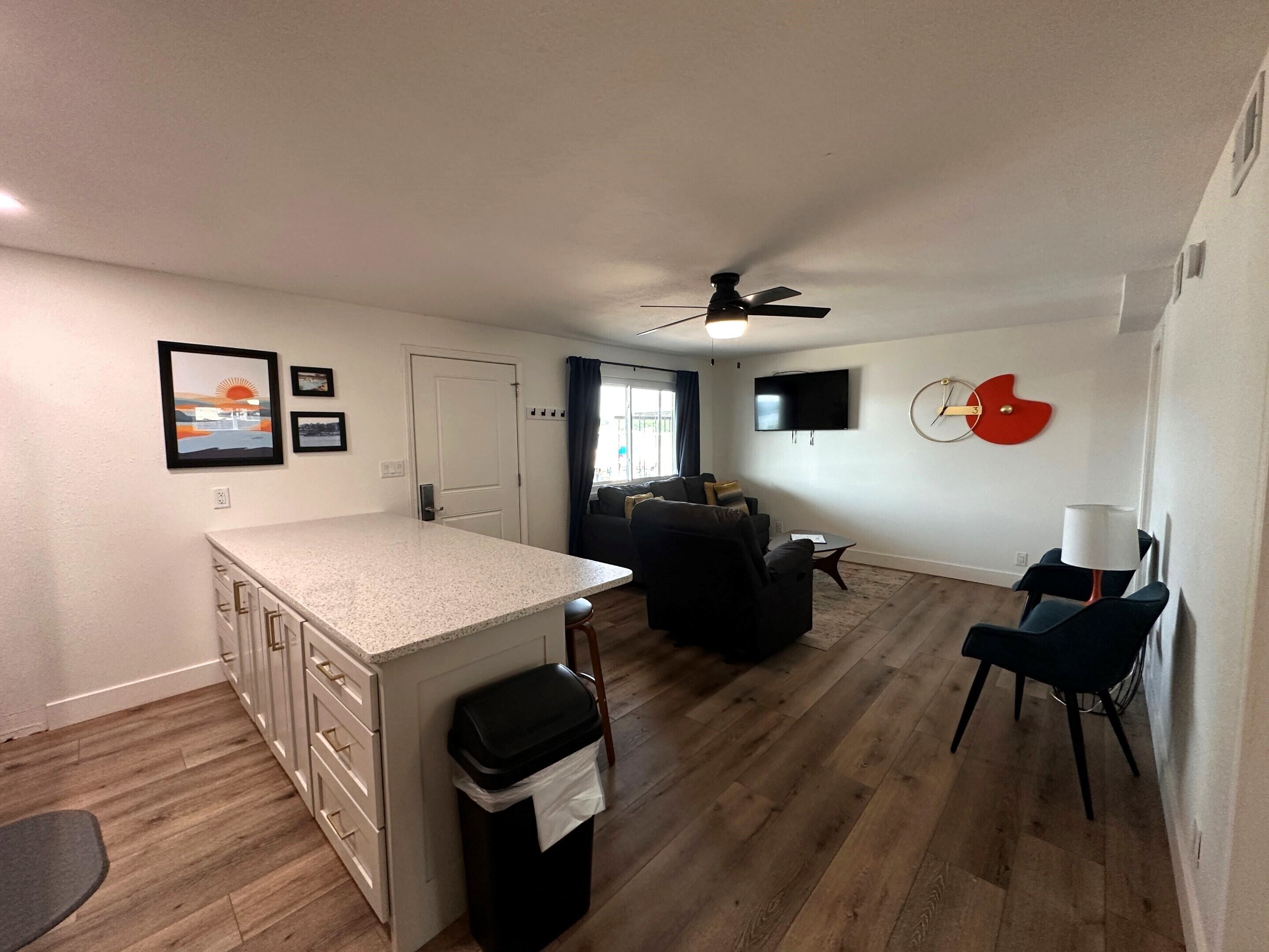A modern kitchen with a central island, a dining area with a high chair, a living room with a sofa, and a TV mounted on the wall.