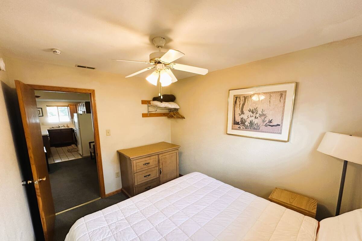 A bedroom with a bed, a ceiling fan, a dresser, a framed picture, and a door leading to a bathroom.
