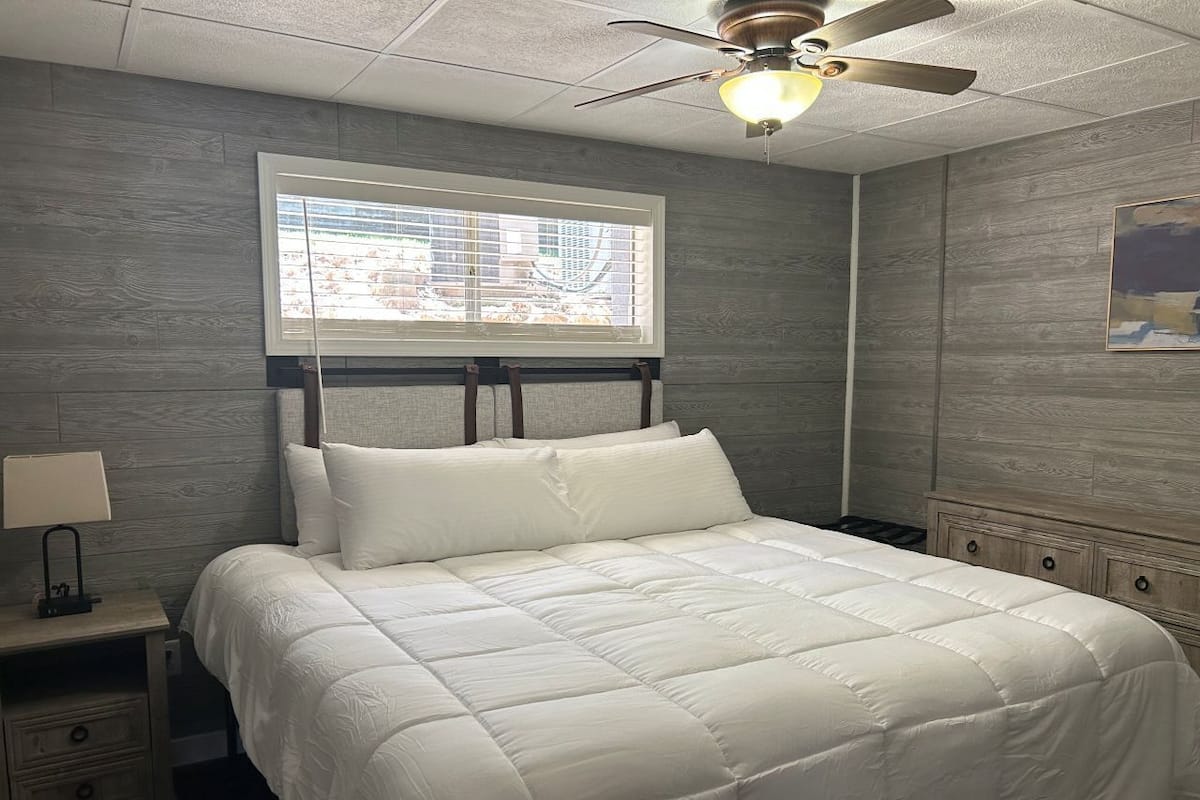 A bedroom with a bed, a nightstand, a lamp, a ceiling fan, and a window with blinds.