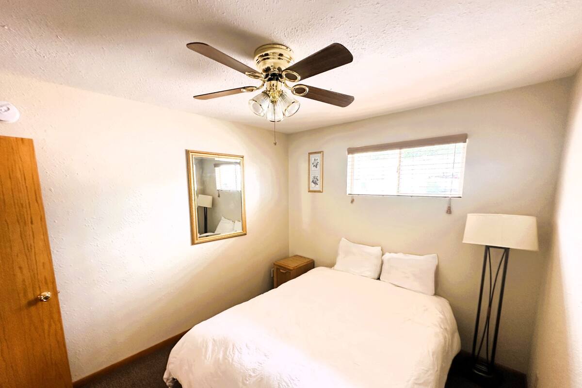 A bedroom with a bed, a ceiling fan, a nightstand, a lamp, a window, and a mirror.
