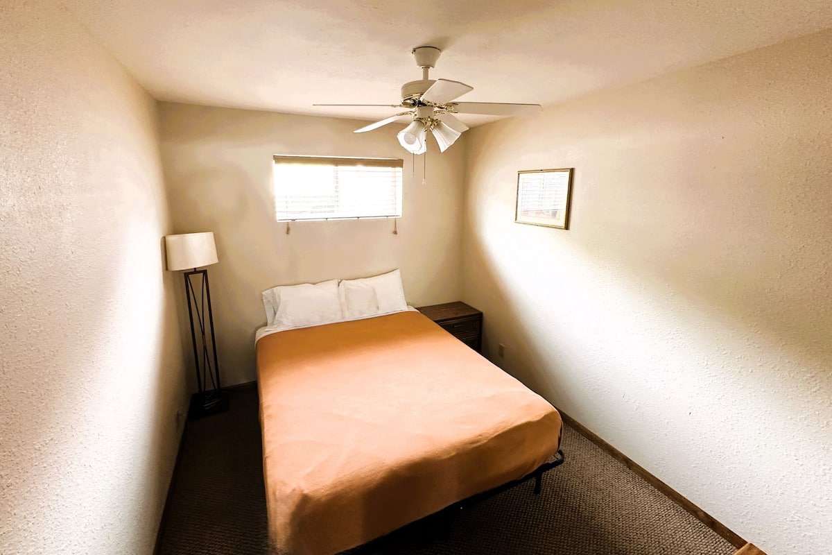 A small, single-bed room with a ceiling fan, a bedside table, a lamp, and a window with blinds.