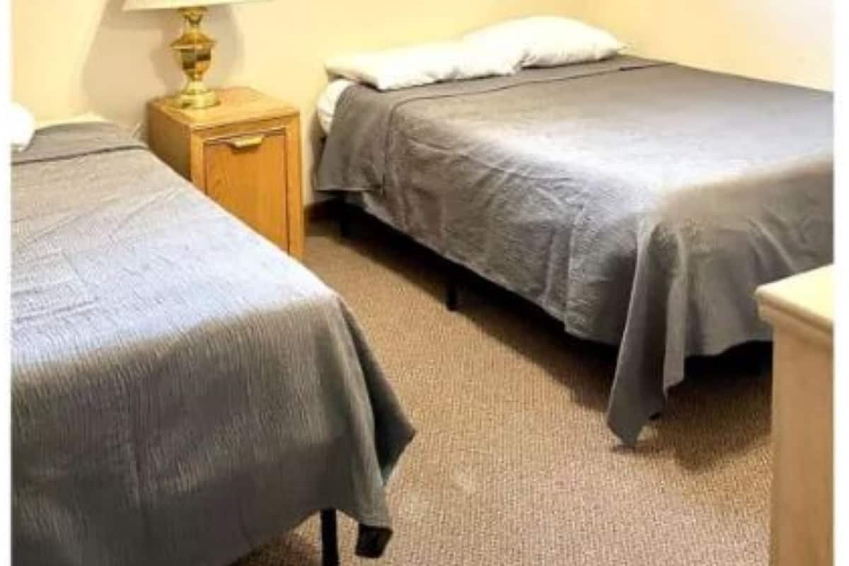 A small, simple room with two single beds, a nightstand with a lamp, and a window with blinds.