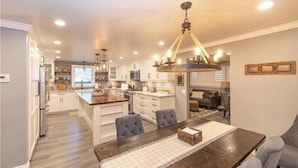 A modern kitchen with a central island, pendant lighting, and a dining area with a wooden table and chairs. - Pine Summit | Hot Tub+Sauna | BBQ | Arcade + Games (Big Bear Lake)
