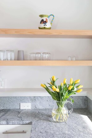 Una encimera de cocina con una jarra de vidrio transparente que contiene tulipanes amarillos, una taza de cerámica con forma de gallo y vasos de vidrio transparente sobre un estante de madera.