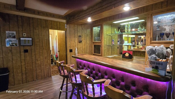 Zona de bar con paneles de madera, barra con encimera iluminada en morado y grifo de cerveza Corona. Hay cuatro taburetes y una caja registradora.