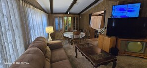 A living room with a brown sofa, a wooden coffee table, a lamp, a dining table with chairs, and a television.