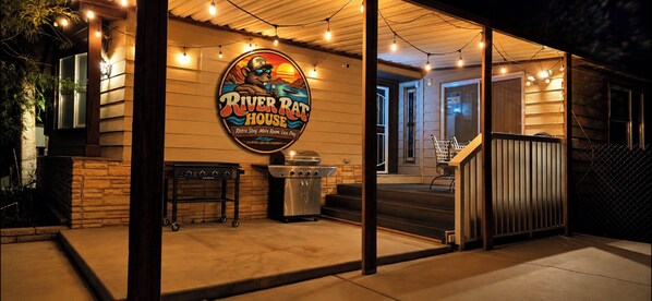 A covered outdoor area with a grill and barbecue equipment, a sign for 'River Rat House,' and a wooden fence.