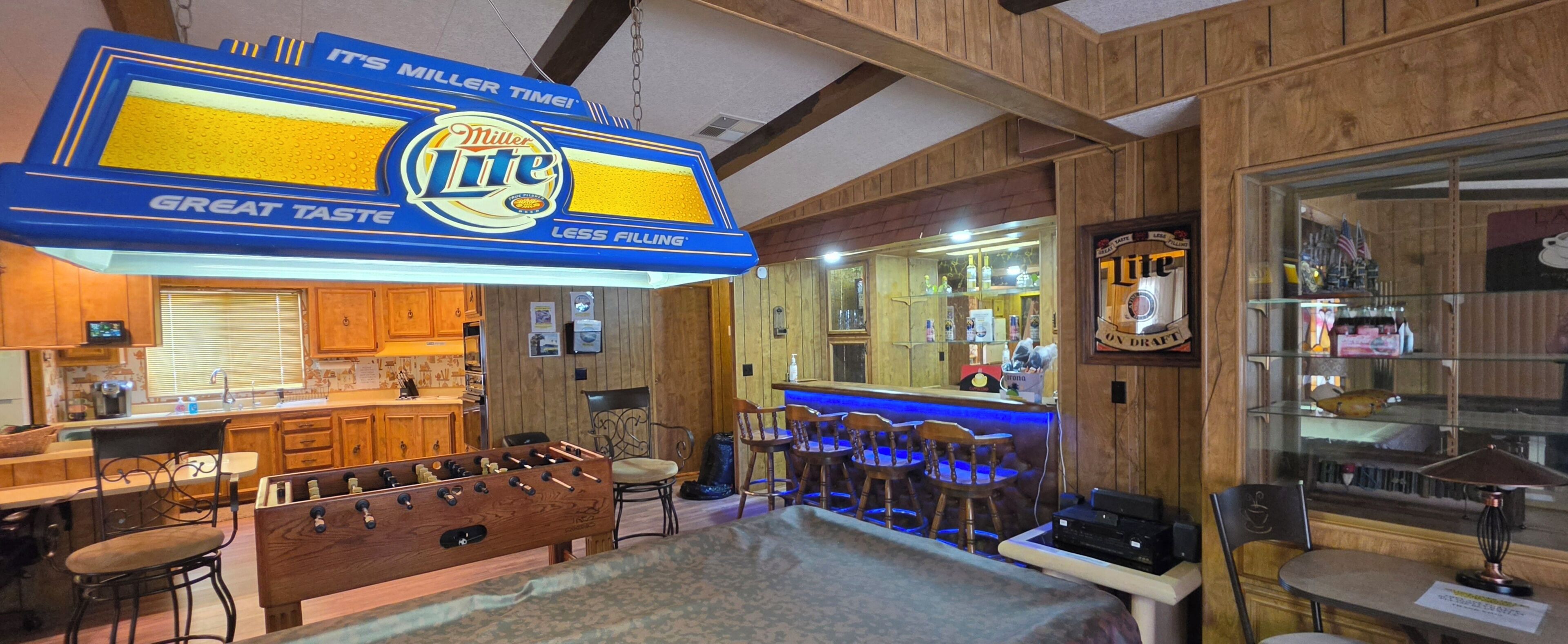 Zona de bar amb un cartell de Miller Lite, tamborets de fusta i una taula de futbolí.
