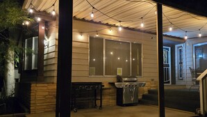 Un espacio al aire libre techado con una parrilla, luces colgantes y vista hacia una casa.