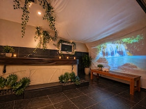 A spa area with a stone bench, potted plants, a fireplace, and a projection of a waterfall.