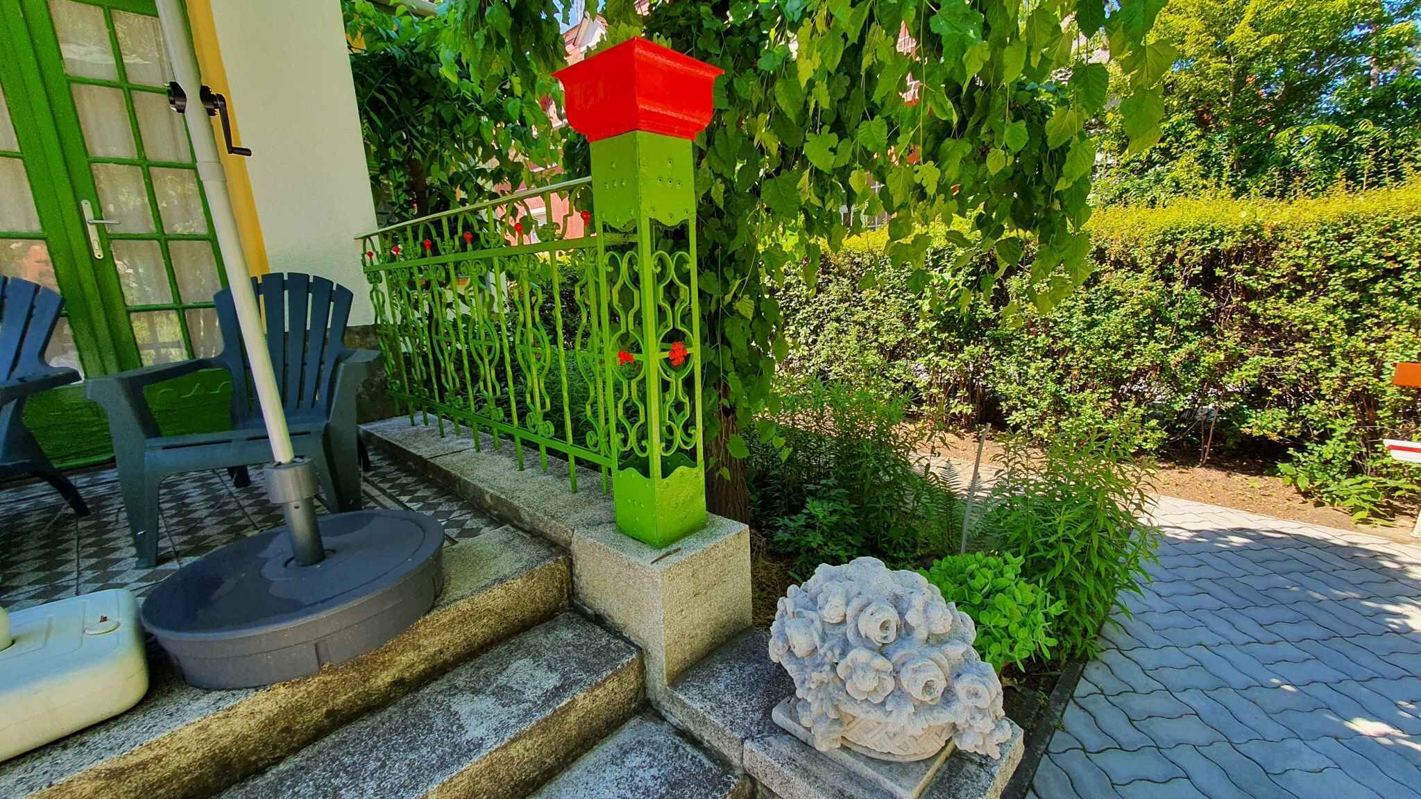 A garden entrance with a green metal fence, a stone sculpture, and a paved pathway.