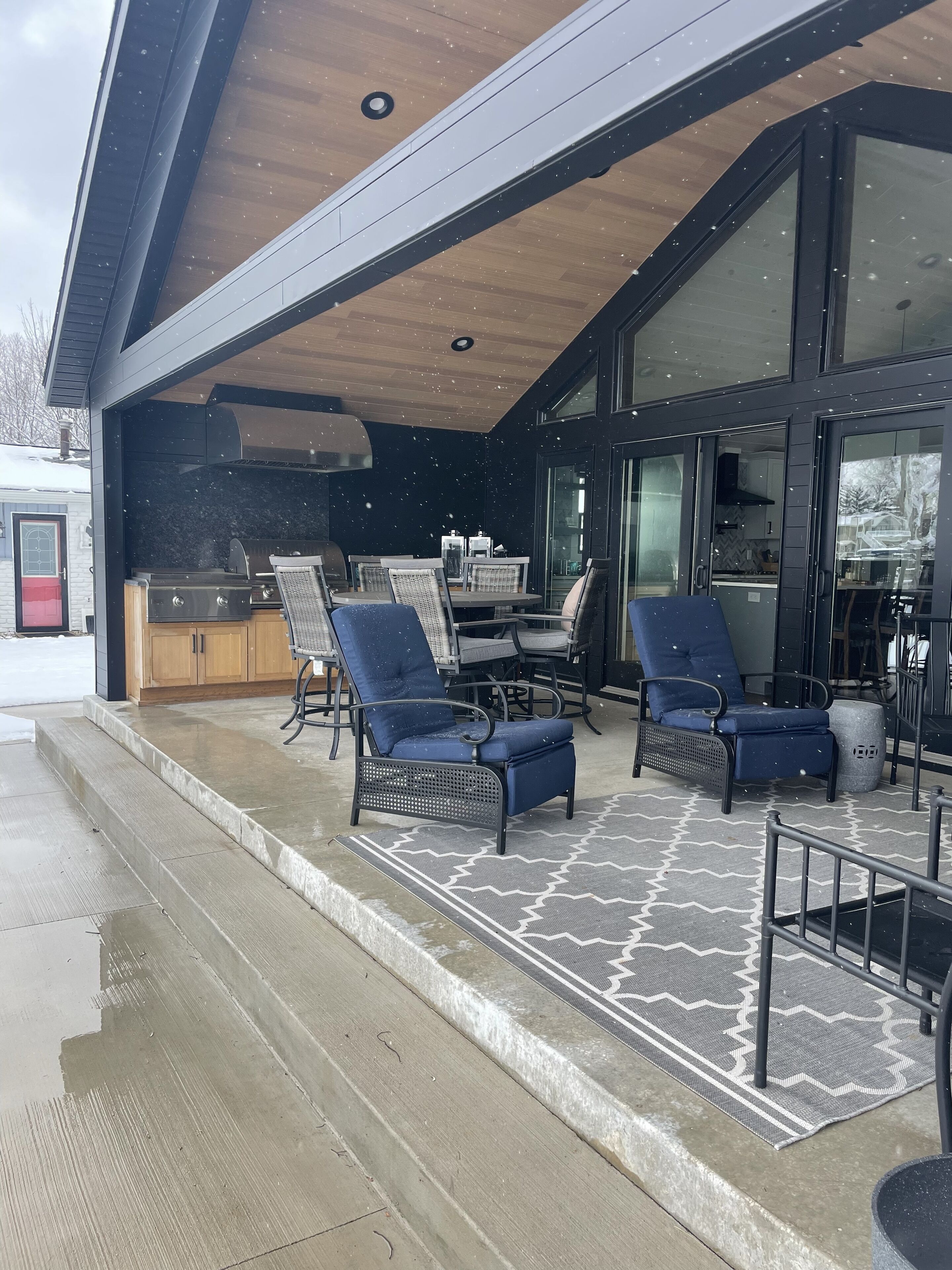 Une terrasse extérieure couverte, équipée d’un barbecue, d’une table à manger et de chaises bleues.